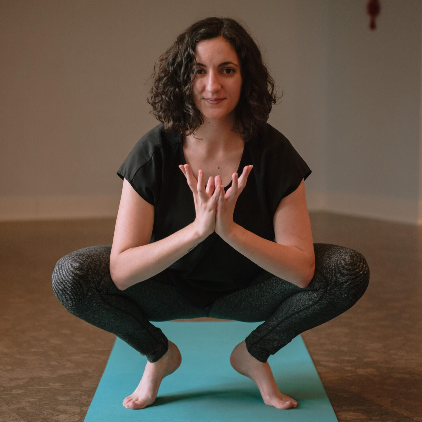 Niki (a white woman with curly brown hair wearing a black shirt and leggings) smiling at the camera in a yogic squat on a teal mat. Her hands are in a lotus mudra.